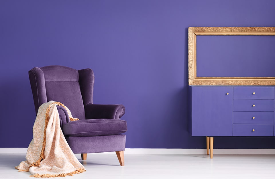 Interno nel colore Pantone 2022, Very Peri: c'è una console con sopra una cornice dorata vuota, e una poltrona viola con una coperta