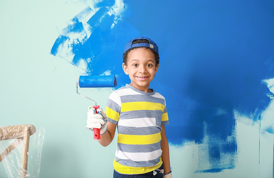 Un bambino con la maglia a righe e un rullo per dipingere intinto di vernice blu, e alle sua spalle una parete parzialmente verniciata