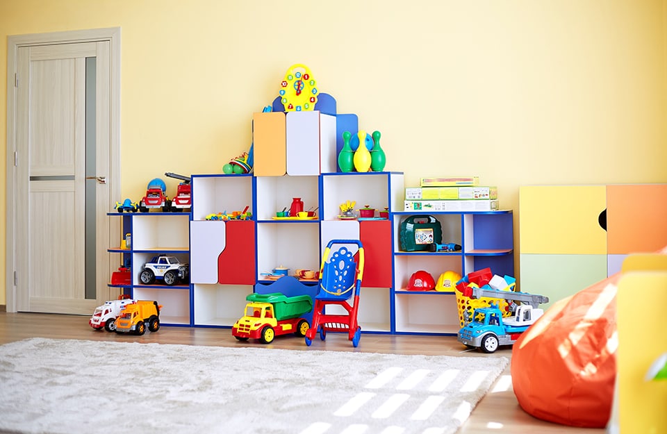 Pared amarilla de una habitación infantil y, junto a la puerta, una gran estantería de colores de forma irregular con juguetes y una gran alfombra de color claro;