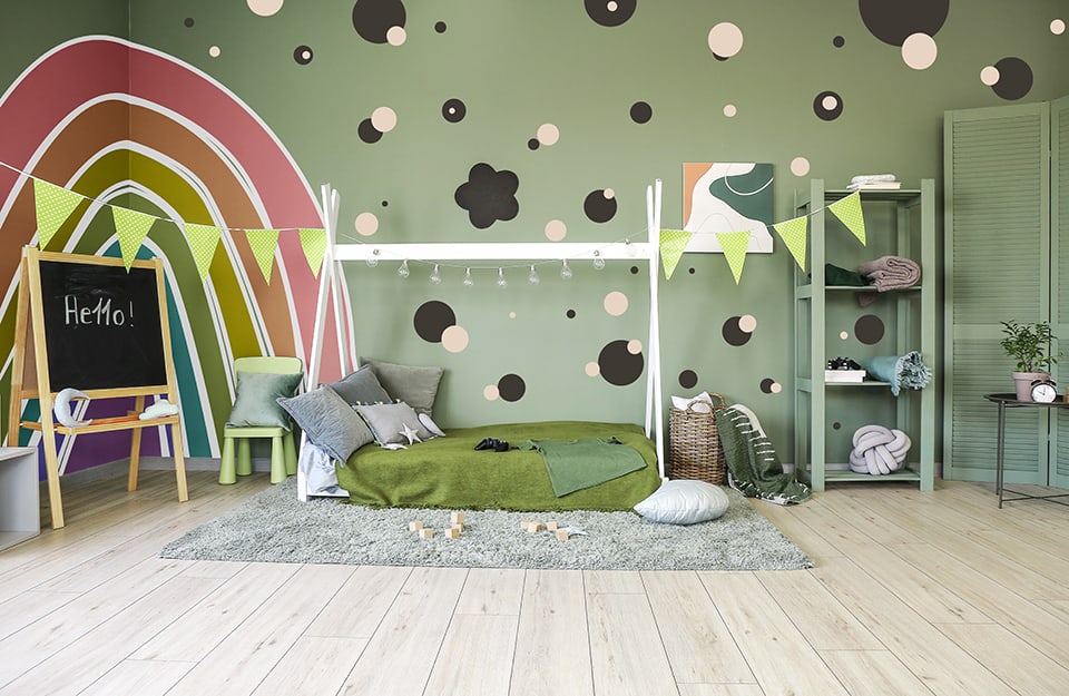 Habitación infantil con una cama blanca con estructura de cortina, paredes verdes decoradas con plantillas, incluido un gran arco iris. Hay una pizarra con 