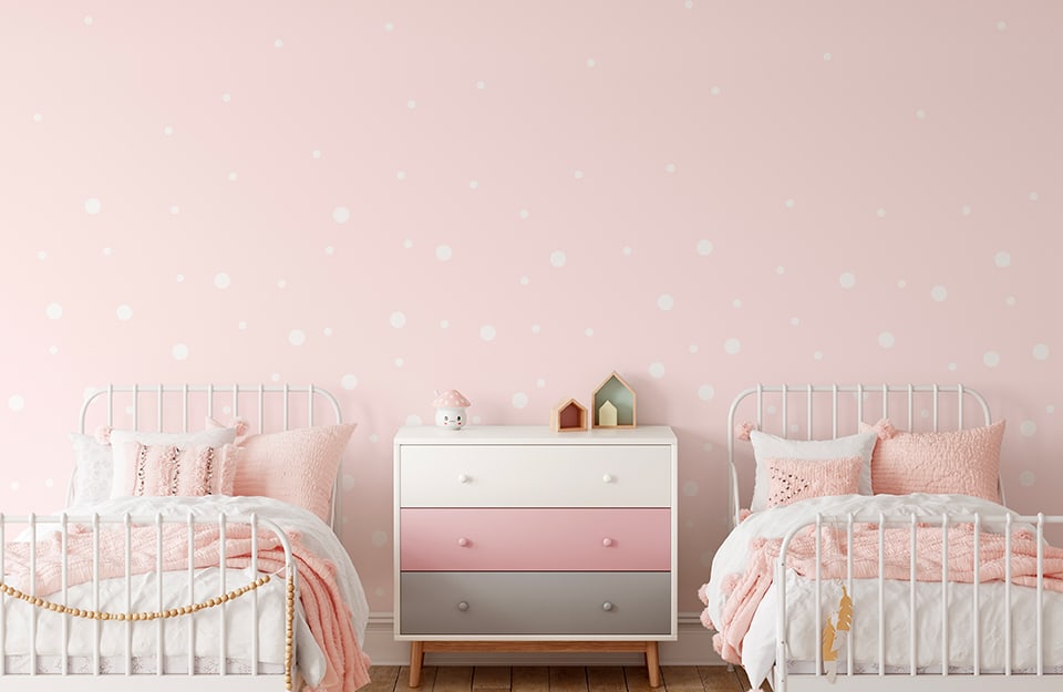 Habitación infantil con dos camas infantiles con armazón de metal blanco. Entre ellas hay una cómoda de distintos colores (blanco, rosa y gris). La pared es rosa, decorada con lunares rosas y blancos, y la ropa de cama es blanca;