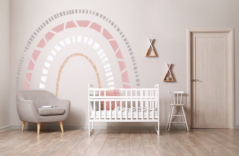 Dormitorio infantil en tonos rosas con un arco iris decorativo en la pared, pintado con signos que parecen arte étnico. Hay una cuna blanca con barandillas laterales, un taburete blanco con respaldo, estanterías en forma de tipi y un sillón gris, además de una puerta;