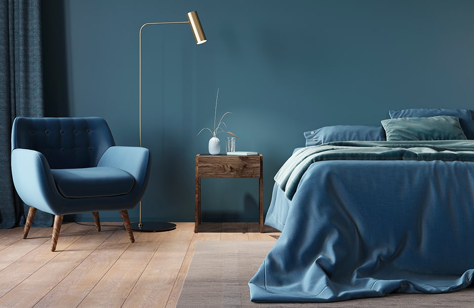 Stanza da letto tutta sui toni del blu, con letto con biancheria blu, parete blu, poltrona blu in stile scandinavo. Il pavimento è un parquet con grandi assi. Il letto è sopra un tappeto grigio-beige e accanto al letto c'è un comodino in legno con sopra un libro, un bicchiere di vetro e un vasetto bianco. Tra la poltrona e il comodino c'è una lampada da terra minimale e dorata