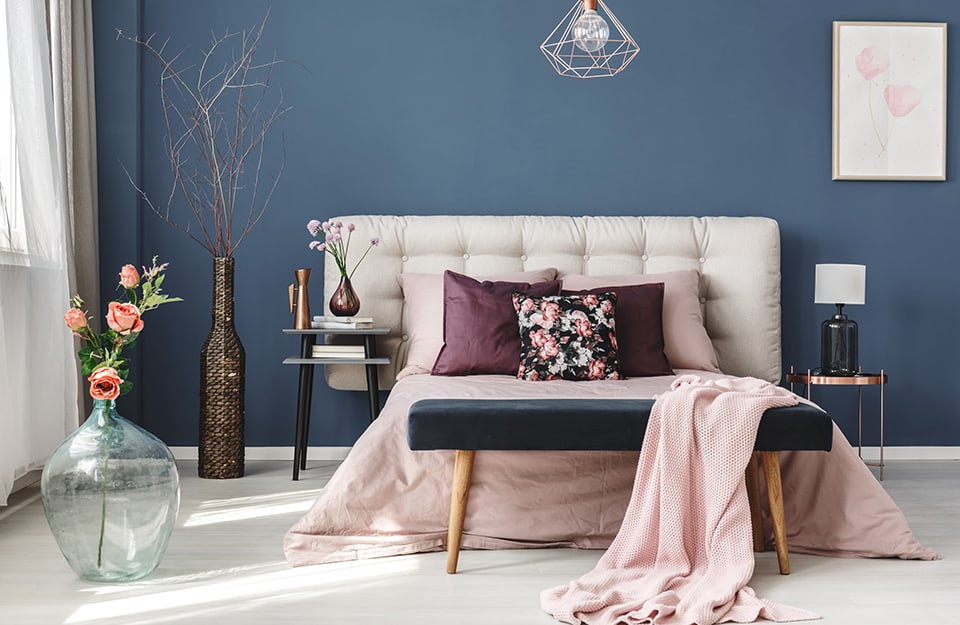 Bedroom with blue wall and white floor. The headboard of the bed is quilted and wider than the bed itself, the two bedside tables are different (wooden on one side, metal on the other). At the end of the bed is a bench with a lined seat, in blue. On the wall is a floral print and from the ceiling comes a chandelier with an LED light and the geometric 'skeleton' of the lampshade. There are various flowers and sprigs in the room, in small or very large vases;