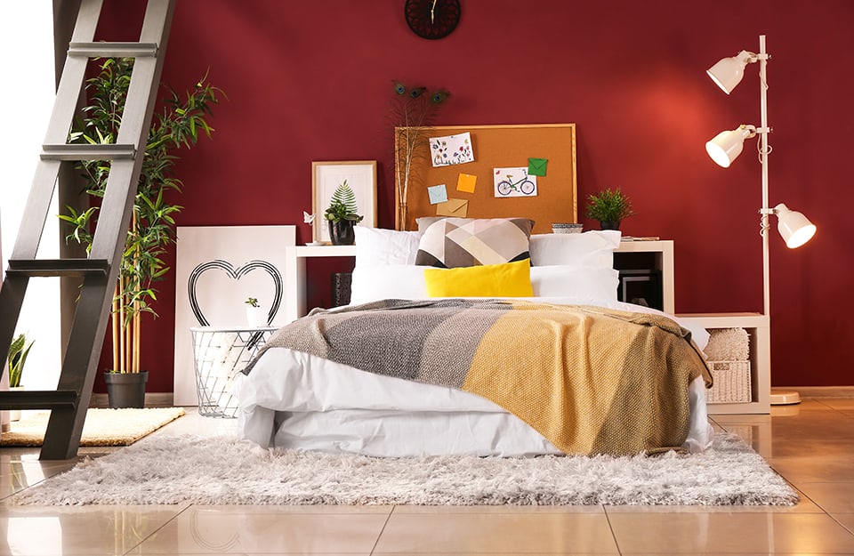 Dormitorio diáfano con paredes rojas. La cama está sobre una alfombra de pelo largo y en lugar del cabecero hay estanterías horizontales, encima de las cuales hay plantas, láminas y un tablero de corcho. Junto a la cama, a la derecha hay una lámpara de pie con tres focos, a la izquierda una mesita, un cuadro y una maceta, mientras que una escalera metálica conduce a un altillo;