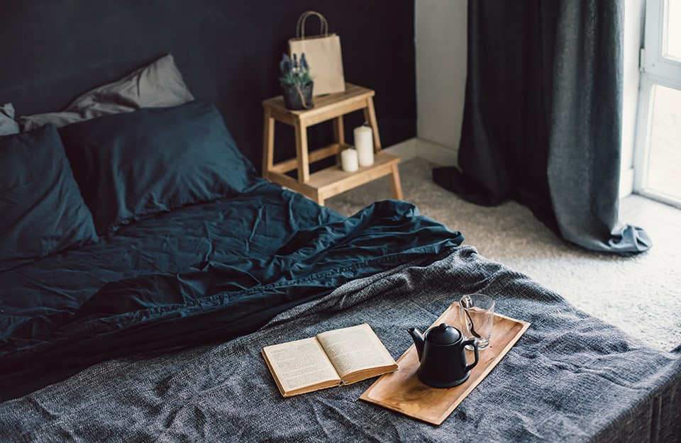 Detalle de una cama de matrimonio con una bandeja con una tetera y un vaso y un libro abierto. La ropa de cama es oscura, el dormitorio tiene las paredes negras y moqueta en el suelo. La mesilla de noche es una escalera/taburete donde hay dos grandes velas blancas, una plantita y una bolsa de papel