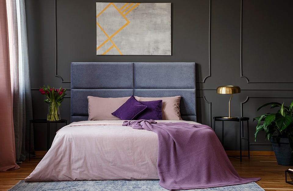 Elegante dormitorio en tonos negros y morados: pared enmarcada en negro, cama de matrimonio con cabecero morado y ropa de cama rosa y morada, alfombra bajo la cama, suelo de parqué, dos mesas auxiliares de metal negro con una lámpara de metal en una y un jarrón con tulipanes rojos en la otra. Encima de la cama hay un cuadro abstracto gris y amarillo y a la derecha se ve parte de una maceta;