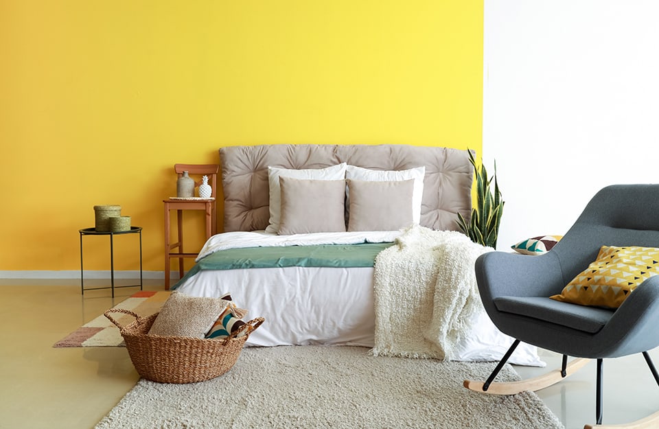 Dormitorio con pared bicolor amarilla y blanca (amarillo a la izquierda y blanco a la derecha). La cama de matrimonio tiene un cabecero tapizado en gris. Hay alfombras a los lados y debajo de la cama, donde también hay una cesta de mimbre con cojines dentro, y una mecedora azul de estilo escandinavo. A un lado de la cama se vislumbra una planta. Al otro, un taburete alto con respaldo, utilizado como mesilla de noche, y una mesita de metal negro con dos jarrones encima. El suelo es claro