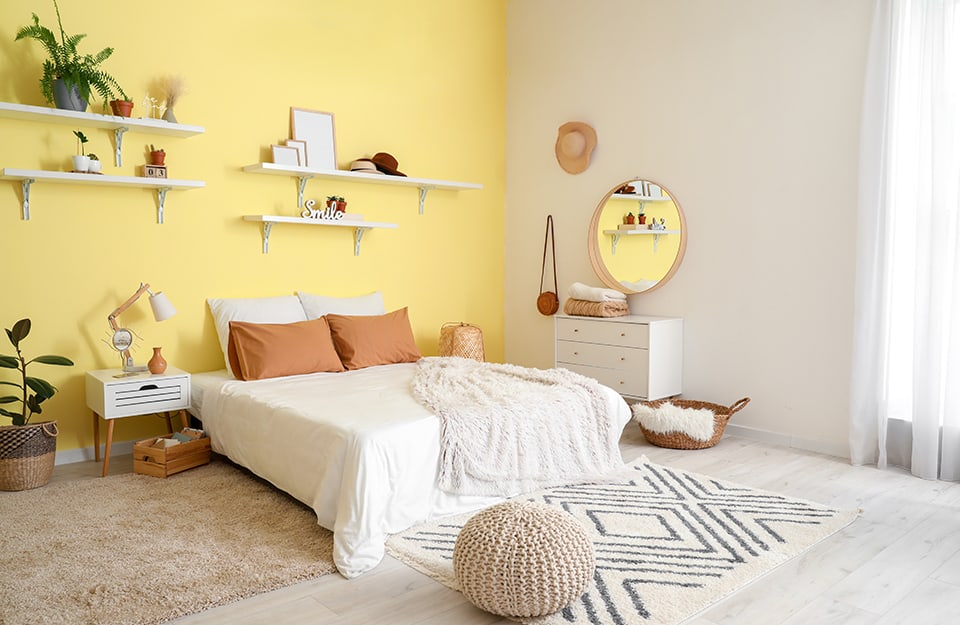 Camera da letto in stile boho-chic sui toni del giallo e del bianco. La parete principale, dietro al letto, è giallo pastello, con diverse mensole che reggono soprammobili, piantine e cornici. Il letto è matrimoniale, con a un lato una lampada in legno intrecciato e all'altro un comodino di legno in stile scandinavo con cassetto e una lampada. In fondo e a un lato del letto ci sono dei tappeti chiari. A terra un parquet bianco. Sulla parete di lato, bianca, ci sono appesi uno specchio circolare in legno, un cappello di paglia e una borsetta in pelle vintage, una cassettiera bianca. A terra un cesto in vimini e un pouff