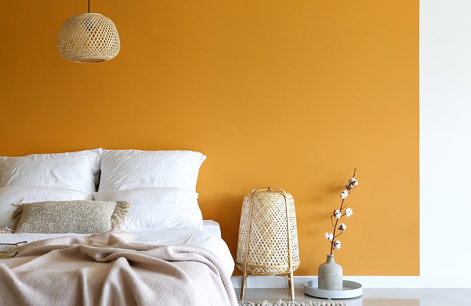 Detalle de un dormitorio con una pared amarillo-naranja con marco blanco. La cama está parcialmente deshecha, con sábanas de color claro. Del techo desciende una pequeña lámpara de araña de madera tejida. Junto a la cama, una lámpara de madera tejida y un jarrón con una ramita en flor en su interior;