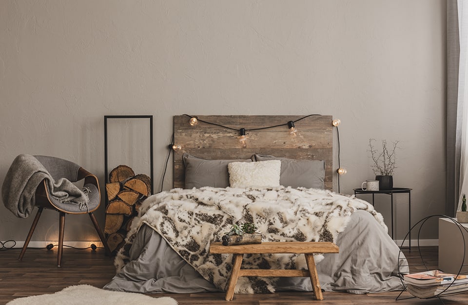 Dormitorio en tonos grises y madera natural. El suelo es de parqué oscuro. La pared es gris. La cama tiene un cabecero de madera vintage recuperada, sobre el que descansa una ristra de lámparas LED. La ropa de cama es blanca y gris y hay una manta de pelo largo que parece piel. Al final de la cama hay un banco vintage de estilo rústico. A un lado de la cama hay una mesa de centro de metal negro con una taza esmaltada en blanco y una jarra negra encima. Al otro lado de la cama hay un marco rectangular de metal con madera de chimenea en su interior. A su lado hay un sillón de estilo escandinavo con asiento gris. En el suelo se ve una alfombra blanca y un revistero de metal esencial;