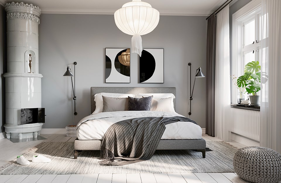 Camera da letto luminosa sui toni del bianco e del grigio. Soffitto bianco, pavimento in parquet bianco, pareti grigie, letto con struttura grigia imbottita, biancheria bianca e grigia, un grande tappeto grigio sotto al letto. Sopra al letto ci sono due stampe astratte geometriche in bianco e nero, e ai lati due lampade in metallo affisse al muro su binari verticali in metallo nero. Sul davanzale della finestra ci sono dei vasi con piante e dalla parte opposta della stanza una grande stufa in ceramica bianca, alta fino al soffitto. Sul tappeto un paio di sneaker bianche e dal soffitto scende un grande lampadario in tessuto bianco