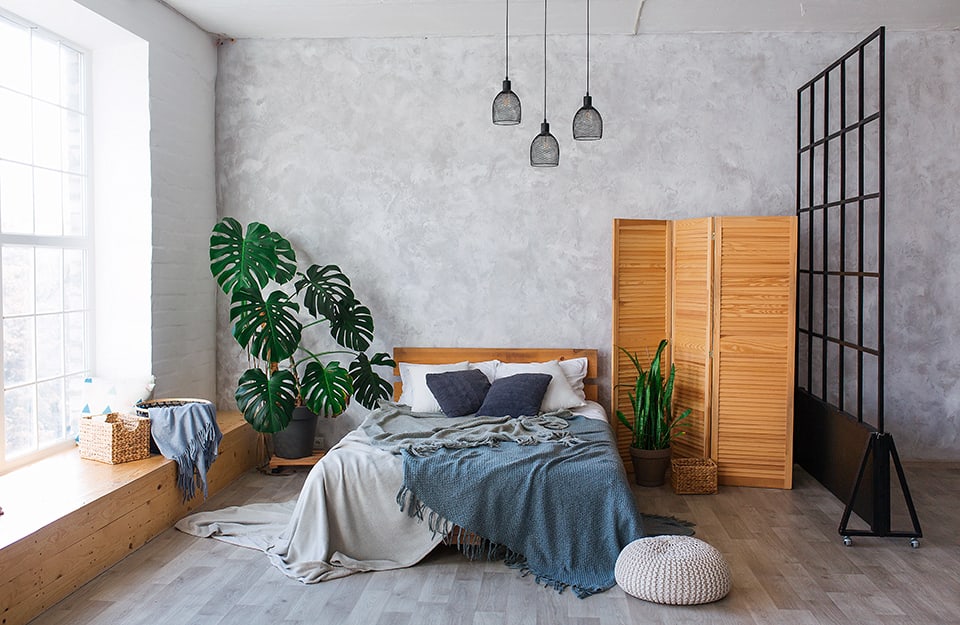 Luminoso dormitorio con suelo de parqué gris y pared de fondo gris claro con efecto arena. Bajo la ventana, un largo marco de madera natural sirve de banco. La cama de matrimonio tiene marco y cabecero de madera. A un lado hay una gran planta (monstera) en una maceta. En el otro, un biombo de madera y una pequeña maceta. Un tabique de metal negro divide el espacio en lo que parece ser un espacio abierto;