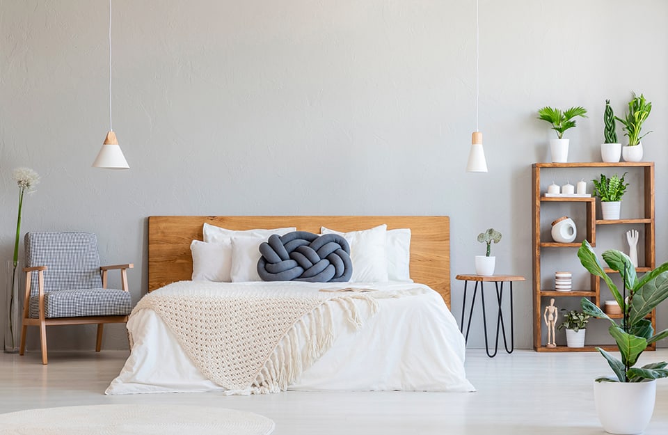 Dormitorio con paredes grises y suelo de parqué blanco. La cama tiene un cabecero de madera natural y ropa de cama blanca. A un lado de la cama hay un sillón de madera con tapicería a cuadros blancos y grises, y un jarrón alto de cristal con una flor grande. Al otro lado de la cama hay una mesita con estructura de metal y plataforma de madera con una maceta encima. A un lado, una estantería de madera con plantas y adornos. A la derecha se ve otra planta en una maceta blanca. A ambos lados de la cama, dos lámparas de araña cónicas de madera blanca y natural descienden del techo, con formas similares pero no idénticas;