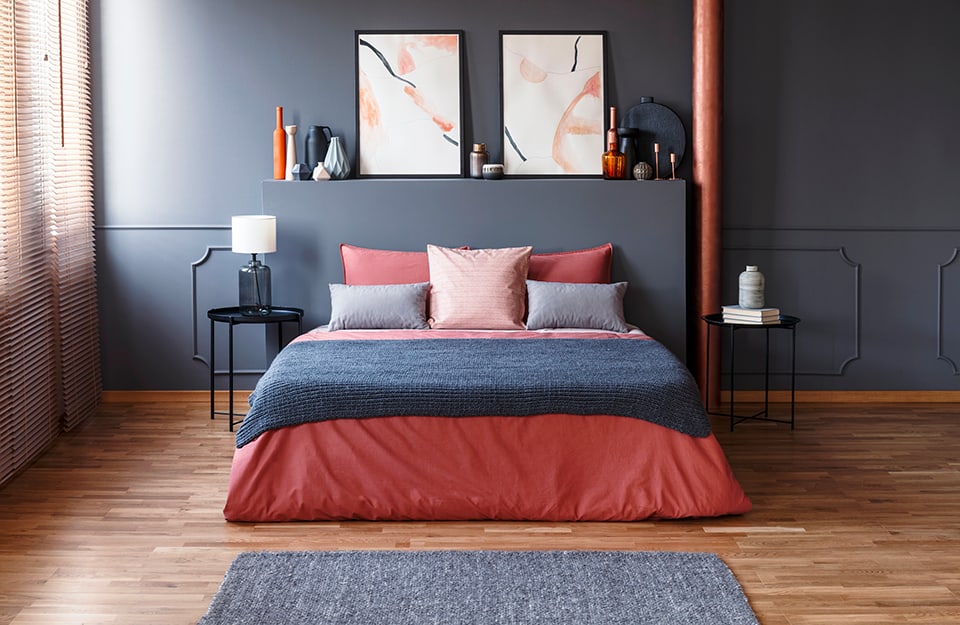 Dormitorio en tonos gris oscuro y rosa. El suelo es de parqué, la cama tiene ropa de cama rosa y gris y la pared del fondo es gris oscuro, decorada con marcos. Detrás de la cama hay un cabecero de escayola tan gris como la pared, y sobre él hay muchos tarros de colores y dos grabados abstractos. A ambos lados de la cama hay dos mesas de metal mínimas, con una lámpara, libros y un tarro encima. A la izquierda hay una ventana con una persiana veneciana;