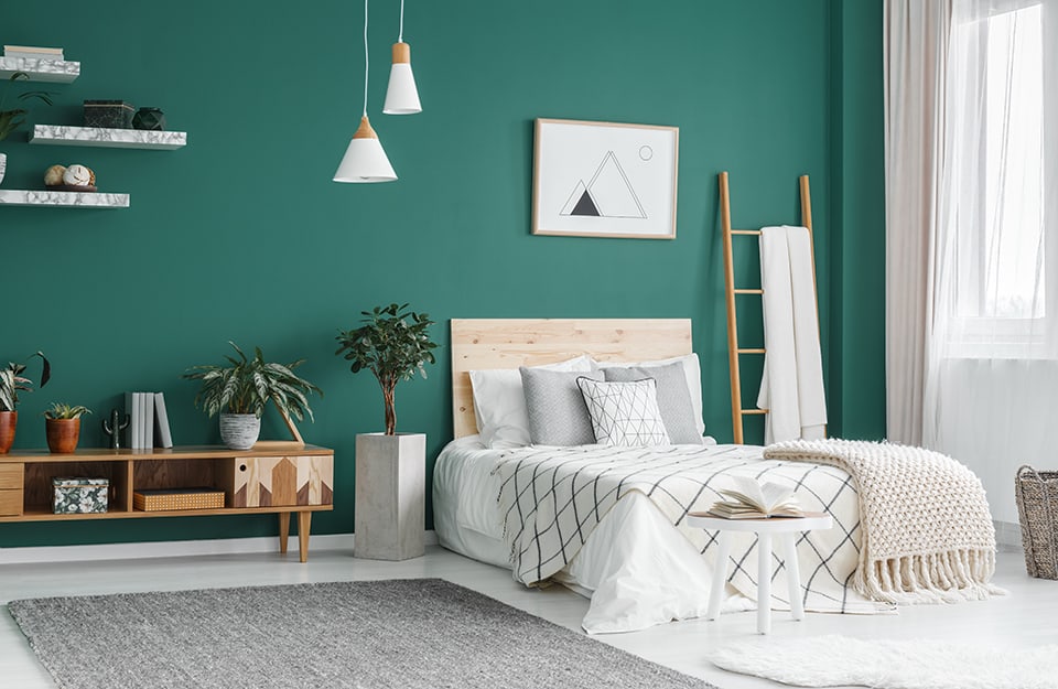 Dormitorio de estilo boho chic con suelo de parqué blanco y pared principal verde intenso. La cama tiene ropa de cama blanca y un cabecero de madera natural clara. A la derecha hay una escalera de madera utilizada como perchero, y hay una ventana cubierta por una cortina blanca semitransparente. Al final de la cama hay una mesita de madera blanca con un libro abierto y una alfombra blanca. Encima de la cama, un estampado con una montaña en estilo minimalista geométrico en blanco y negro. Del techo descienden dos lámparas de araña cónicas blancas similares pero de formas diferentes. A la izquierda de la cama hay un jarrón de hormigón en forma de paralelepípedo con una planta en su interior, y una estantería horizontal de madera de estilo modernista. Encima, fijadas a la pared, tres estanterías blancas efecto mármol con chucherías