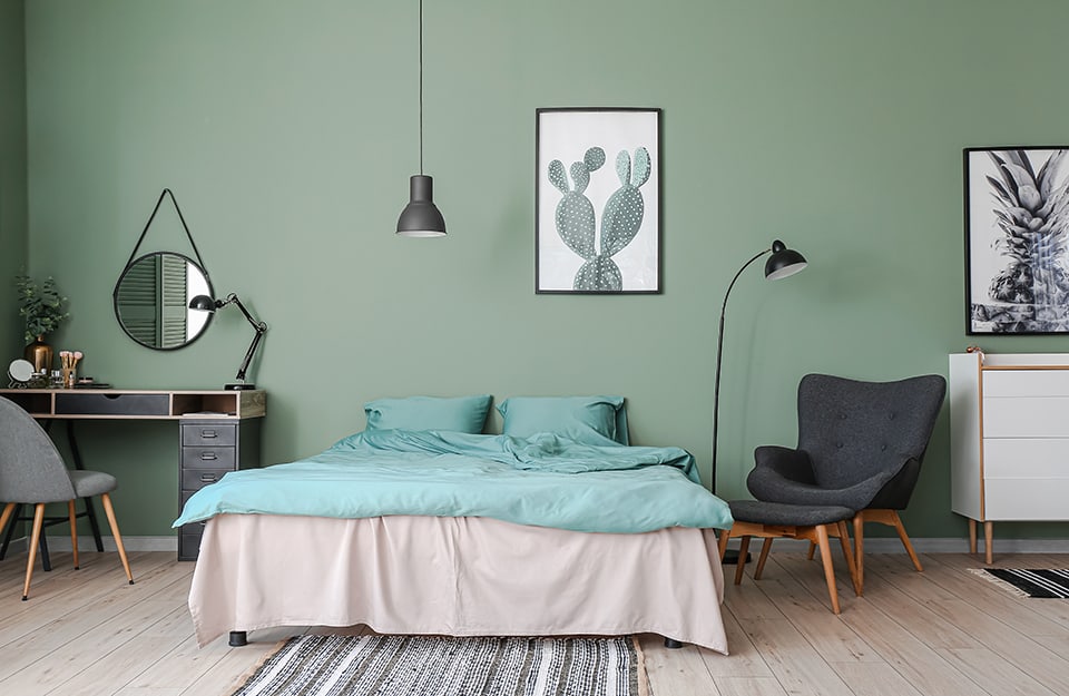 Ampia camera da letto con pareti verdi e pavimento in parquet chiaro. Il letto è matrimoniale, con biancheria grigia e verde acqua. Sopra al letto una stampa botanica. A un lato del letto, una lampada da terra in metallo nero, una poltona grigia in stile scandinavo con poggiapiedi abbinato e separato, e un mobiletto bianco a cassetti con sopra una foto in bianco e nero a tema botanico. Dall'altra parte del letto, una toletta essenziale con cassetti, in legno e nera, con sedia e, appeso al muro, uno specchio circolare con cornice nera