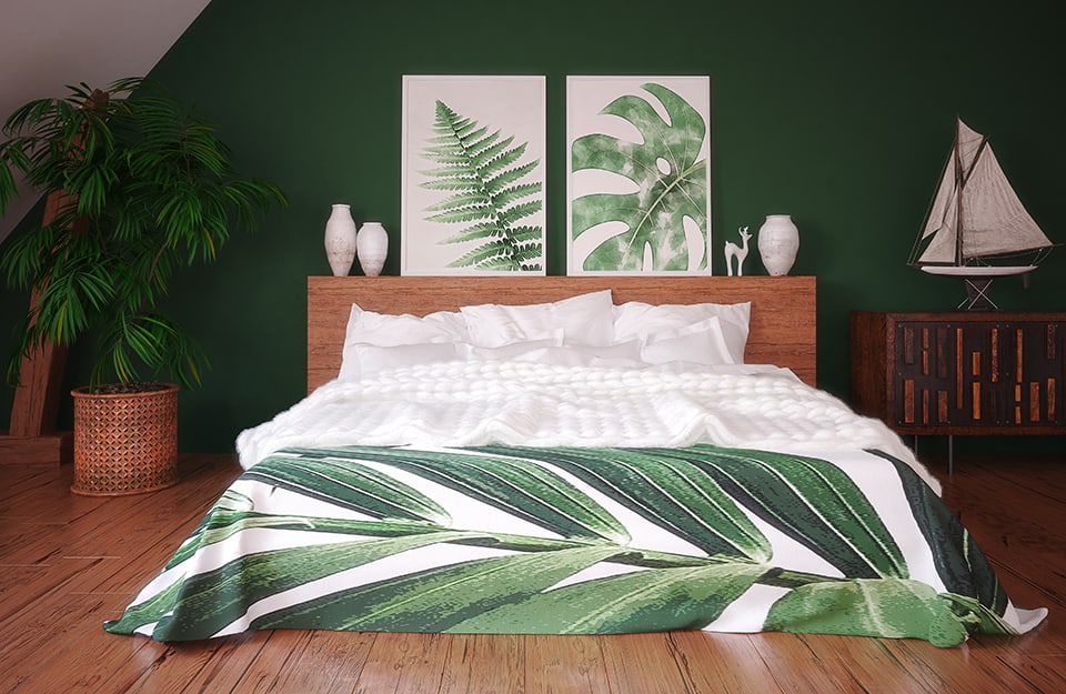 Habitación abuhardillada con dormitorio. El suelo es de parqué y la pared del fondo, verde. La cama tiene un marco de madera, con un gran cabecero, sobre el que hay jarrones blancos y dos estampados de temática botánica. La manta también es de temática botánica. A ambos lados de la cama hay una gran planta en un lado y en el otro un armario de madera de estilo étnico con una maqueta de un velero encima;