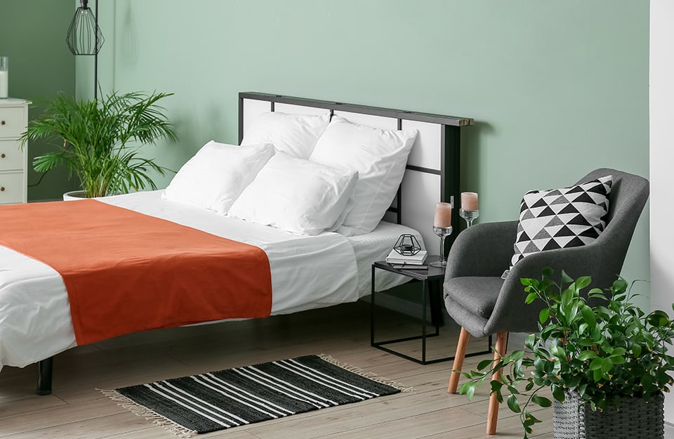 Dormitorio con vistas de tres cuartos, con paredes verdes, suelo de parqué, cama de estructura de madera con cabecero blanco y negro. A ambos lados de la cama, por un lado, una mesa esencial de metal negro con libros, velas y tarros sobre ella, y un pequeño sillón gris de estilo escandinavo, así como una planta en una maceta de mimbre gris y una pequeña alfombra de rayas blancas y negras. En el otro, una planta en una maceta blanca y un mueble de cajones también en blanco;