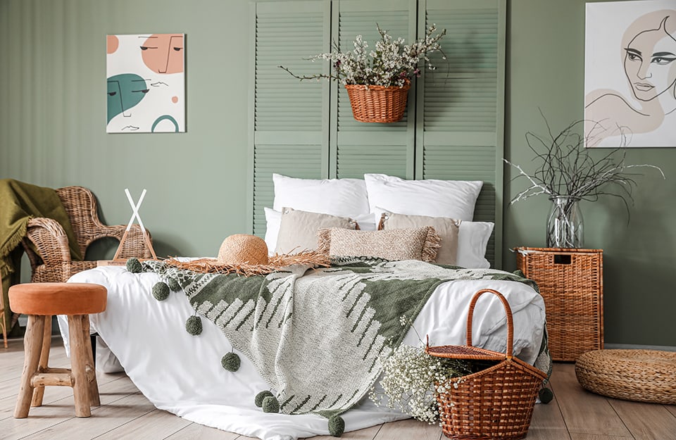 Camera da letto in stile rustico moderno, sui toni del legno e del verde. Parete verde, testiera del letto della stesso colore, realizzate con vecchie persiane. Pavimento in parquet, coperte in cotone a motivi geometrici con ponpom e cappello di paglia sul letto. Nella stanza ci sono diversi elementi in vimini (una poltrona, due cesti, un comodino con sopra un vaso, e molti fiori e rametti. Alla parete sono appesi due quadri con volti stilizzati. In fondo al letto c'è uno sgabello rustico con seduta rosa