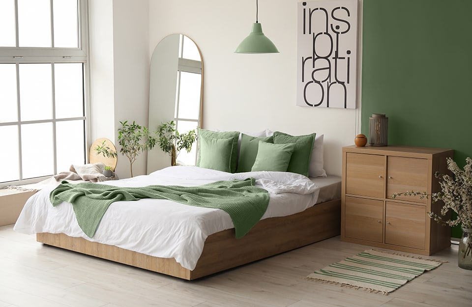 Camera da letto moderna in stile essenziale in grande stanza con ampia finestra a tutta altezza. Il letto ha una struttura in legno squadrata ed essenziale, con biancheria verde e bianca. La parete dietro al letto è bianca e a un lato, verso la finestra, è appoggiato uno specchio ad arco, dietro una sedia piena di vestiti. Sopra il letto c'è una stampa grafica con scritto “inspiration” e inizia poi una parete verde, alla quale si appoggia in armadietto in legno squadrato con quattro sportelli, davanti al quale c'è un piccolo tappeto a motivi geometrici. Sopra il letto scende, dal soffitto, un lampadario verde come la parete