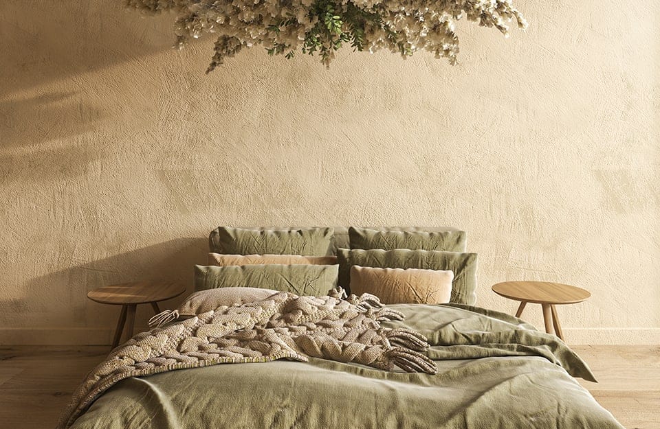 Dormitorio de estilo mediterráneo con una pared beige de efecto arenado. La cama es de tonos verde pastel, y a ambos lados hay dos pequeñas mesas circulares que hacen las veces de mesillas de noche. Desde arriba, se vislumbra una composición floral que baja del techo, iluminada desde la derecha