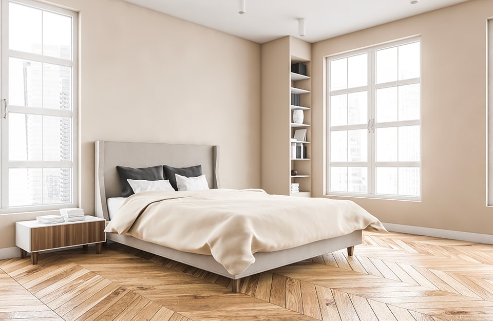 A bright and spacious bedroom in an essential style, with beige walls, a bed with sober lines, beige bed linen, herringbone parquet flooring, a low, square bedside table in white and wood, and a plasterboard bookcase in the same colour as the wall, rising to the ceiling. Two large windows illuminate the room