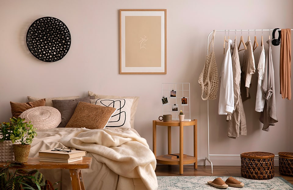 Dormitorio en tonos beige, lleno de elementos. La pared es beige, con un estampado mínimo y un círculo de mimbre pintado de negro. La cama está llena de almohadas y una manta, en tonos pastel de beige y marrón. Al final de la cama hay un espartano banco de madera, desgastado por el tiempo, con libros, unas gafas y plantas sobre él. A un lado de la cama, una mesita redonda de madera con una taza, un tarro y una rejilla de fotos encima. Aún a la derecha, un tendedero de metal blanco con varias prendas y accesorios colgados. Debajo, una alfombra, cestas de mimbre y un par de zapatillas;