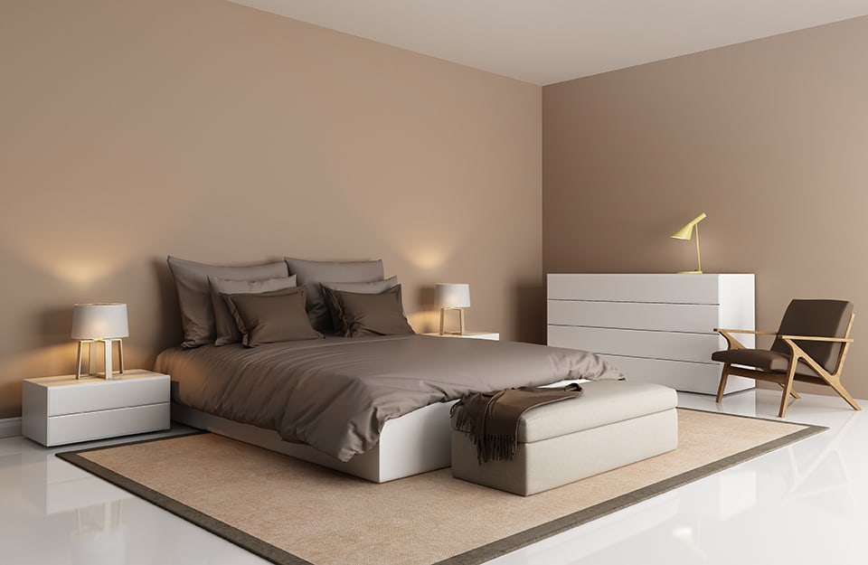 Un dormitorio minimalista en tonos beige, con paredes beige, techo blanco y suelo blanco. Vemos la habitación en vista de tres cuartos, con un somier blanco cuadrado y ropa de cama marrón. A ambos lados de la cama hay dos mesillas blancas cuadradas con cajones y lámparas encima. En una pared, una cómoda del mismo estilo que las mesillas, con otra lámpara encima. Junto a la cómoda, una silla de madera con asiento de cuero marrón, de estilo escandinavo. Debajo de la cama una gran alfombra beige y marrón y al fondo un banco tapizado beige;