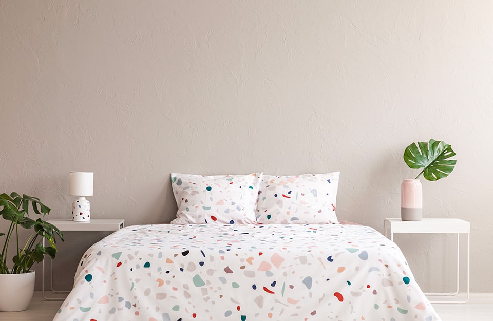 Camera da letto minimal con parete beige, letto con coperta e cuscini bianchi con elementi colorati stile terrazzo, motivo che si ripete anche sulla lampada da notte, che si trova su uno dei due comodini, entrambi in metallo bianco e minimali. Sull'altro comodino c'è un vaso semplice bicolore grigio e rosa, con dentro la foglia di una monstera. Una pianta in vaso appare sulla sinistra