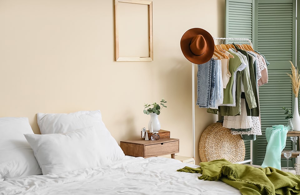 Dormitorio con pared blanca marfil. Se ve una cama con sábanas blancas y una manta verde. Al lado hay una mesilla de noche de madera de estilo modernista con un cajón. Encima hay varios tarros. Colgado de la pared hay un marco vacío de madera clara y apoyado en la pared hay un biombo verde. Entre la cama y el tabique hay un tendedero blanco con varias prendas colgadas y un sombrero marrón. Un poco más atrás, una mesa-escalera con algunos jarrones apoyados en ella;