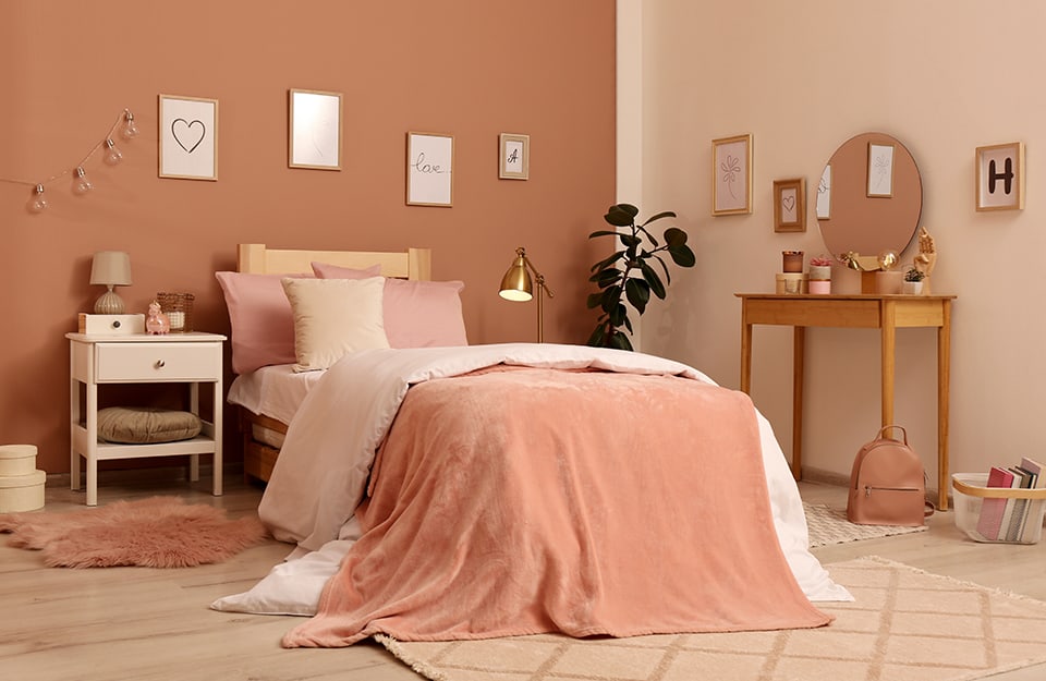 Camera da letto sui toni pastello del beige e del cipria. Si vede una parete sul nocciola e una sul beige, con appesi molti quadretti con scritte e lettere dell'alfabeto. Il letto è singolo, con struttura semplice, in legno, e molte coperte sui toni del cipria, del rosa e del beige. Accanto al letto c'è un comodino in stile rustico dipinto di bianco, con sopra oggetti e una lampada. Ai lati del letto tre diversi tappeti (uno peloso, e due a pelo corto). Dal lato del letto opposto al comodino c'è una pianta. Poggiata alla parete c'è una toletta in legno con molti oggetti sopra e uno specchio circolare. A terra una borsa e un cestino portariviste