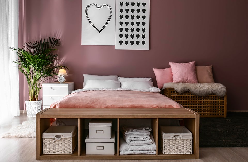 Un dormitorio moderno de estilo romántico con una pared rosa oscuro y suelo de parqué. Al final de la cama hay una estantería horizontal baja con cajas de cartón y ropa de cama. A un lado de la cama hay una mesilla de noche blanca con cajones y una gran maceta. Al otro lado hay un banco de madera tejida con cojines rosas. Encima de la cama hay dos láminas con corazones dibujados en negro