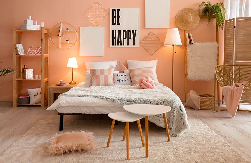 Camera da letto in stile rustico-romantico, con pareti rosa e parquet chiaro. Sotto al letto c'è un grande tappeto color panna. Il letto ha una semplice struttura in metallo nero e ci sono molti cuscini rosa e con scritte. In fondo al letto un cuscino peloso rosa e una coppia di tavolini bianchi dalle forme irregolari. Dietro al letto delle stampe e una griglia metallica con piccole luci a led. Sulla stessa parete, a sinistra del letto, un tavolino semplice in legno con sopra una lampada, una mensola metallica circolare appesa al muro, e una semplice libreria in legno con diversi oggetti in esposizione. Dall'altro lato, una lampada da terra, una scala a piola che regge un tappeto peloso e dei libri. C'è un cappello di paglia appeso, e un separè in legno, oltre a una sedia in bambù