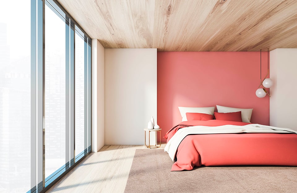 Grande stanza da letto con una parete interamente finestrata. L'arredamento è minimale, con soffitto e parquet in legno, pareti bianche e una parete rientrante in rosa in cui si trova il letto, con biancheria bianca e rosa. Sotto al letto c'è un grande tappeto beige. A un lato del letto un comodino metallico di forma circolare con sopra vasi bianchi. Dal soffitto scendono due lampade bianche sferiche