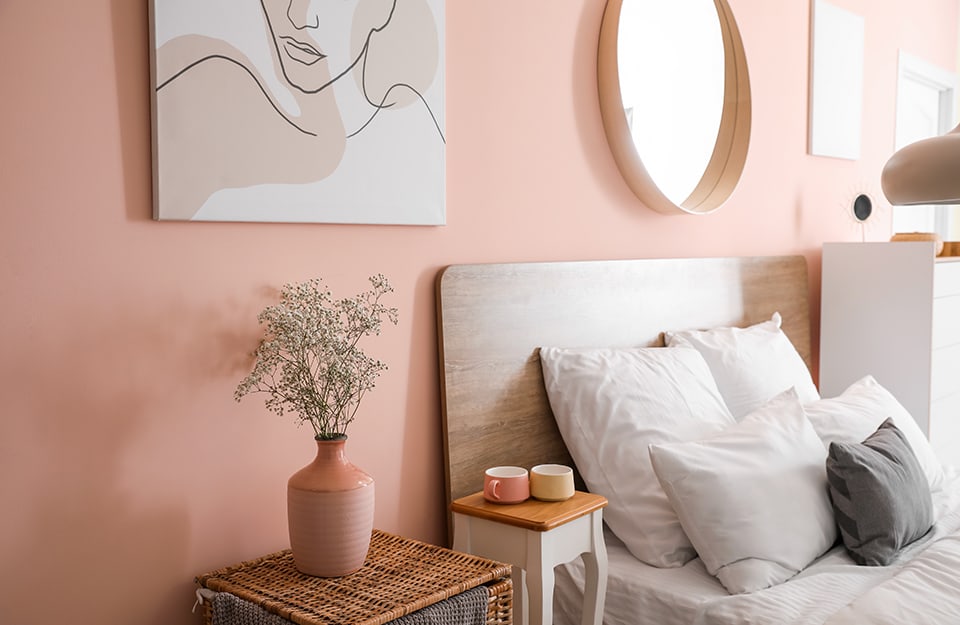 Detalle de un dormitorio con una pared rosa con cuadros colgados y un espejo circular sobre la cama. El cabecero es fino, de madera y con líneas minimalistas. Junto a la cama hay una mesita de madera con tazas y otra de mimbre con un jarrón rosa con flores. En el lado opuesto de la cama hay una cómoda alta y blanca;