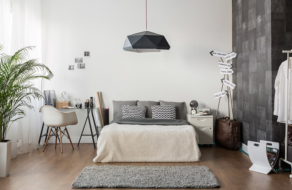 Camera da letto moderna con parquet. Un letto matrimoniale con cuscini grigi davanti a una parete bianca con appese delle fotografie senza cornice. Da soffitto scende un grande lampadario grigio dalla forma sfaccettata. A sinistra del letto c'è una scrivania su cavalletti neri, con una sedie moderna bianca. Sulla scrivania oggetti da ufficio e soprammobili. A destra del letto, un comodino bianco con cassetti, una vecchia valigia di pelle e un appendiabiti in metallo con attaccati cartelli di indicazione delle distanze con i nomi di diverse città (Oslo, Wien, New York, ecc.). C'è anche un portariviste bianco e una parete tappezzata con carta da parati grigia separa la stanza da un altro ambiente