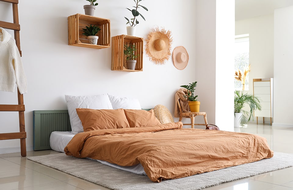 Dormitorio de estilo boho-chic con paredes blancas, una cama apoyada directamente en el suelo, encima de una alfombra y detrás de una vieja contraventana pintada de verde que hace de cabecero. A un lado de la cama hay una escalera de madera que sirve de perchero. Al otro lado hay una vieja silla de peluche sobre la que hay ropa y una planta en una maceta amarilla. Encima de la cama hay cajas de fruta que sirven de estanterías y contienen plantas. Colgados de la pared hay también dos sombreros de paja. Más allá de una columna se vislumbra otra habitación;