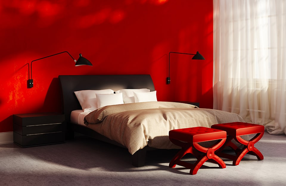 Stanza da letto con pareti rosso intenso e pavimento grigio. Una tenda semi-trasparente bianca copre la finestra. Al muro sono appese due lampade nere direzionabili e ai piedi del letto ci sono due sgabelli rossi trapuntati e laccati