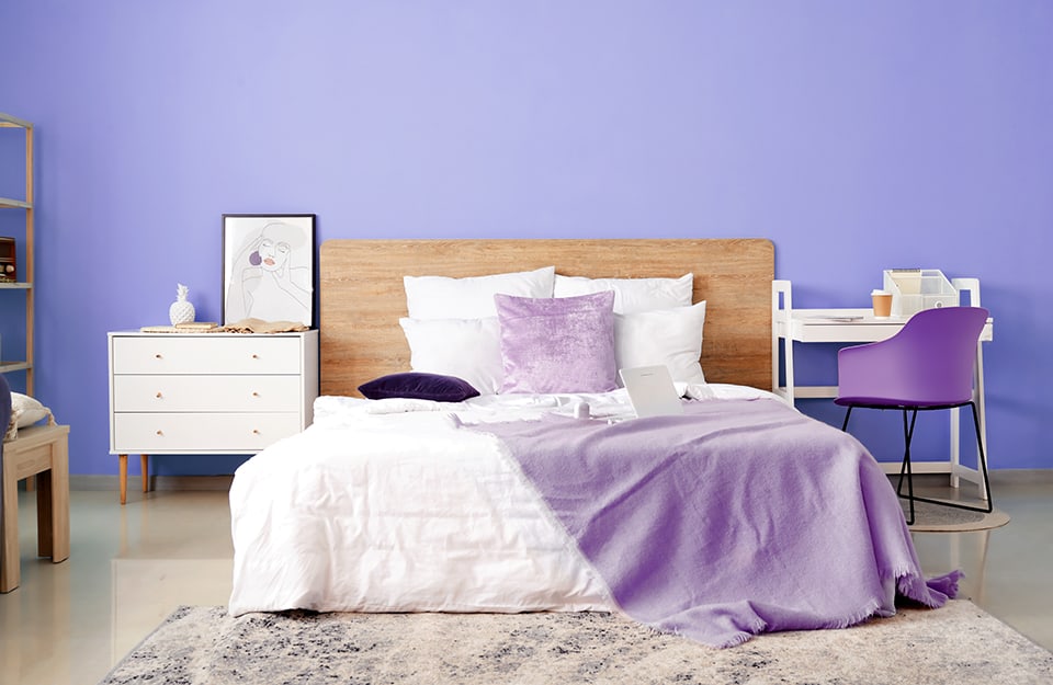 Dormitorio con pared de color púrpura bígaro. La cama tiene un sencillo marco de madera con un gran cabecero. Las almohadas y las mantas son de color blanco y rosa. A los pies de la cama hay una alfombra gris de pelo largo. A un lado de la cama hay un pequeño escritorio blanco minimalista con una moderna silla morada con estructura de metal negro y asiento de plástico. Al otro lado hay una cómoda con patas, y sobre ella un libro, objetos y un grabado con un retrato femenino. Aún a la izquierda, se vislumbran una mesita y una librería;