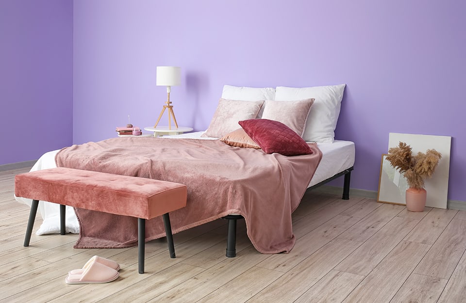 Camera da letto molto spoglia, con parete viola. Il letto è su una semplice struttura in metallo nero. Coperte e cuscino sono sul bianco e sul rosa. A un lato del letto un tavolino circolare bianco con sopra una lampada e un altro tavolino più piccolo con libri e candele. Dall'altro lato, delle cornici poggiate al muro e un vaso rosa con dentro fiori secchi. Il pavimento è in parquet chiaro e si vedono delle ciabatte rosa