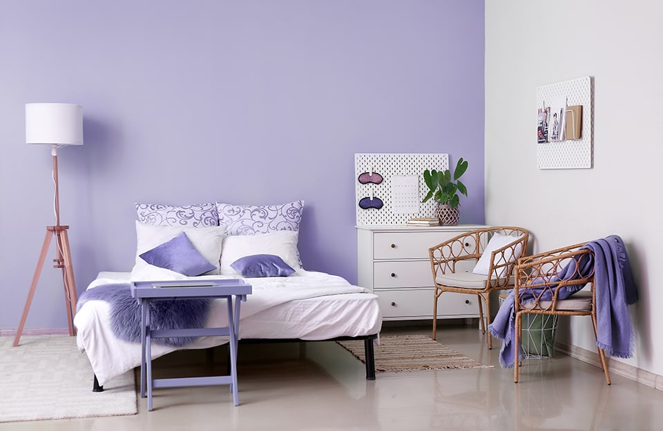 Camera da letto in stile moderno e minimale con parete viola chiaro. Il letto è su una semplice struttura metallica nera. Le coperte e i cuscini sono sul bianco e sul viola. A un lato del letto, sopra un tappeto bianco, una lampada con treppiedi in legno. In fondo al letto un tavolino viola. Dall'altro lato del letto un tappeto più piccolo, una cassettiera bianca con sopra libri e una pianta e due sedie in bambù. Alle pareti sono appesi due pannelli portaoggetti bianchi: su uno ci sono libri e stampe, sull'altro delle mascherine da notte e un calendario