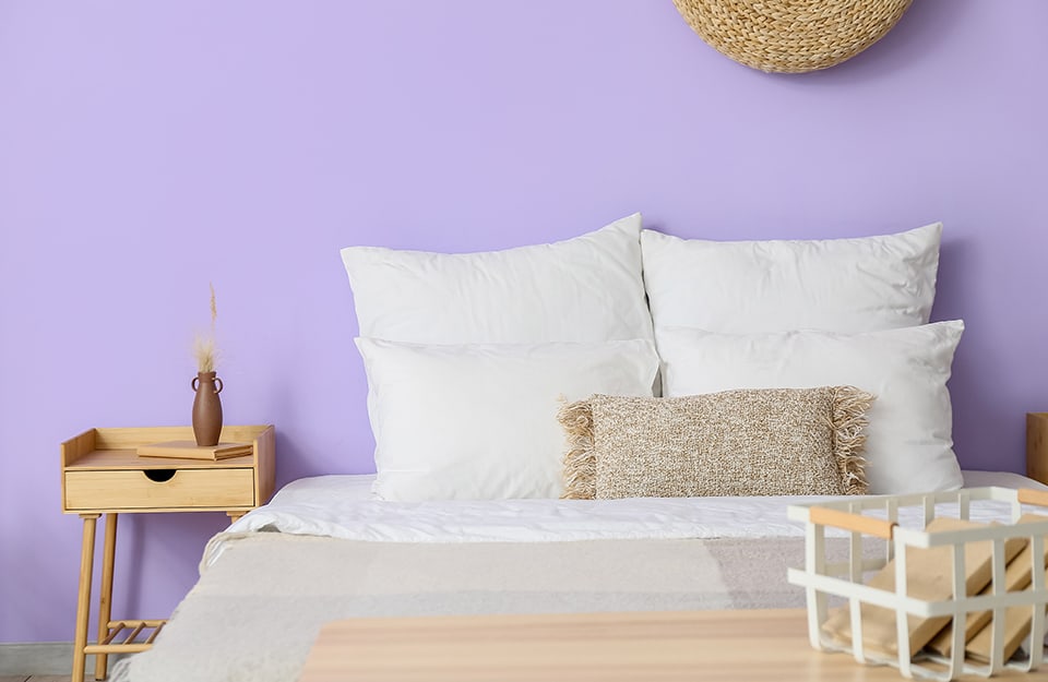 Detalle de un dormitorio con una pared morada . Junto a la cama hay una mesilla de noche de madera de estilo escandinavo, con un libro y un tarro encima. En la pared se vislumbra un colgante circular de mimbre. Al final de la cama hay un banco de madera con una cesta encima en la que hay libros;