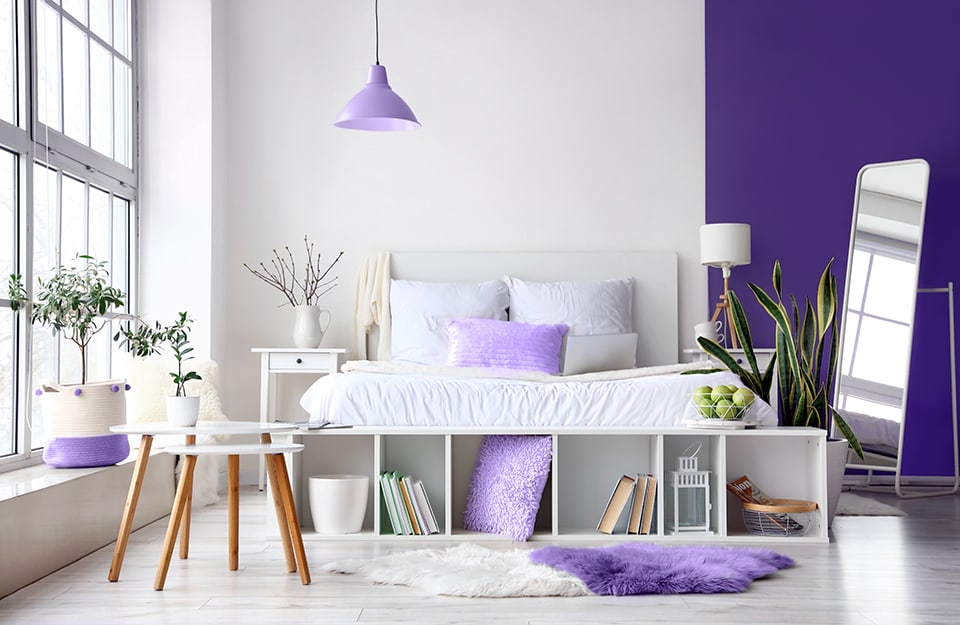 Luminosa e spaziosa camera con grande finestra e davanzale con appoggiato un vaso bianco e viola con una pianta. Il parquet è bianco. La parete dietro al letto è bianca e poi diventa viola. Il letto è in legno verniciato di bianco e in fondo c'è una scaffale basso orizzontale che contiene libri e un cuscino viola. Ci sono altri cuscini viola nella stanza, e sul pavimento c'è un tappeto lungo bicolore bianco e viola. Ai due lati del letto ci sono dei comodini bianchi con un cassetto, e sopra una lampada e un vaso. Dal soffitto scende un lampadario viola in stile industriale. Alla destra del letto c'è una pianta in vaso bianco. Poco oltre un grande con struttura in metallo