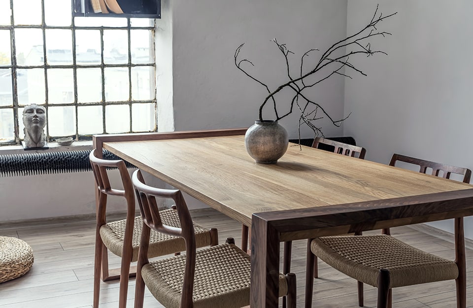 A wooden dining table in a minimalist style, with a metallic vase containing twigs on top. Illuminating the scene is an industrial window with a sculpture of a human face resting on the windowsill;