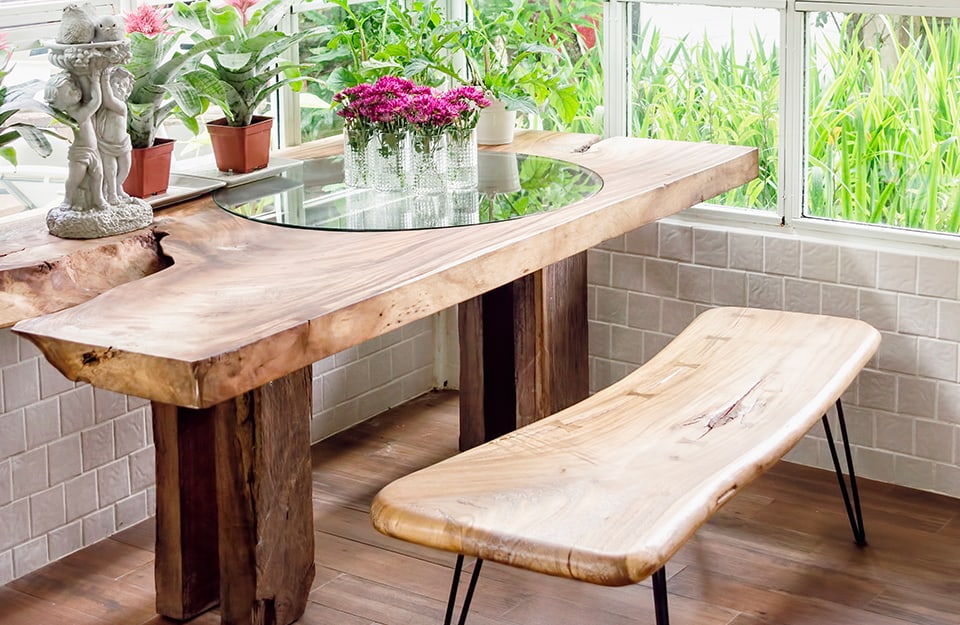 Mesa y banco de estilo rústico y líneas irregulares, con plantas y estatuillas encima, en la esquina de una habitación con ventana esquinera, parquet y paredes de azulejos;