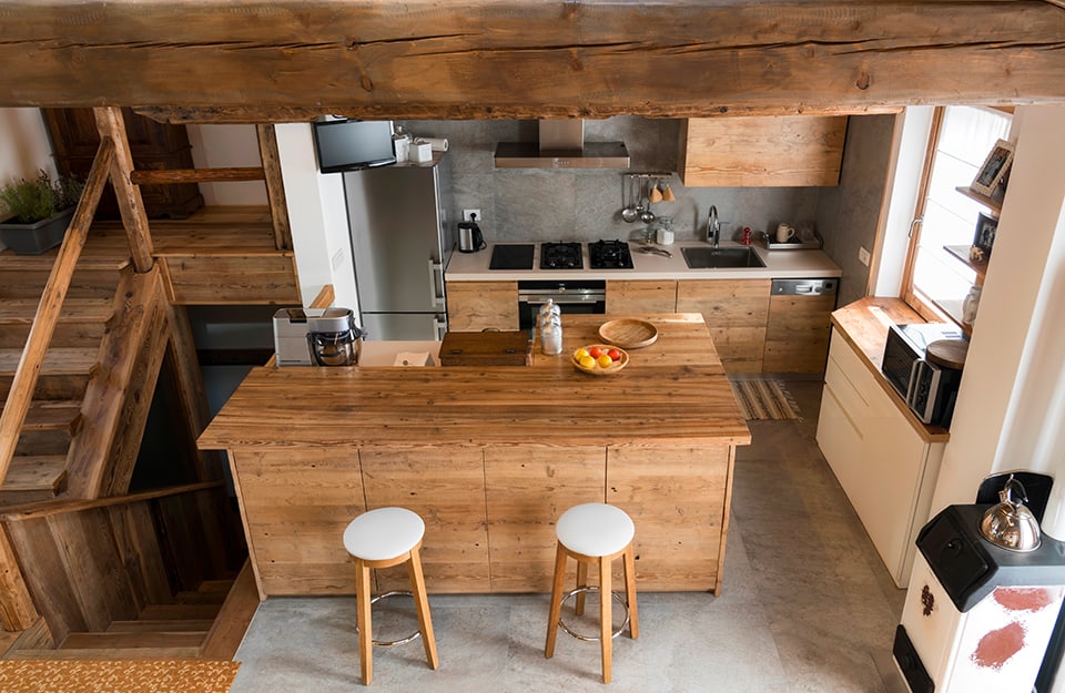 Vista desde arriba de una cocina de estilo rústico con vigas de madera, muebles de madera natural, isla con taburetes y doble escalera que sube y baja a la izquierda