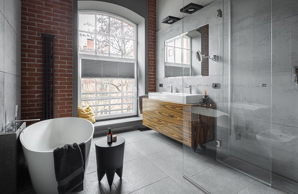 Cuarto de baño de estilo industrial en una habitación con paredes de hormigón rugoso y otra pared con ladrillos vistos. La ducha es totalmente transparente y hay una bañera blanca de estilo minimalista. El mueble-lavabo es de madera natural con vetas muy evidentes