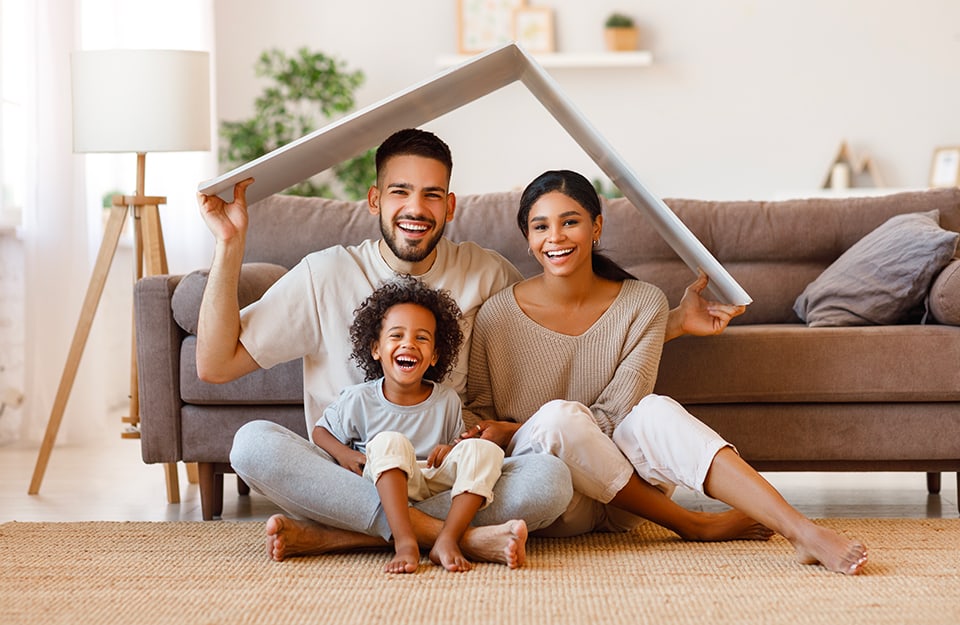 Una famiglia formata da padre, madre e figlia, tutti sorridenti, sorridono mentre, nel loro salotto, tengono sopra alle loro teste un pezzo di cartone bianco a imitare un tetto