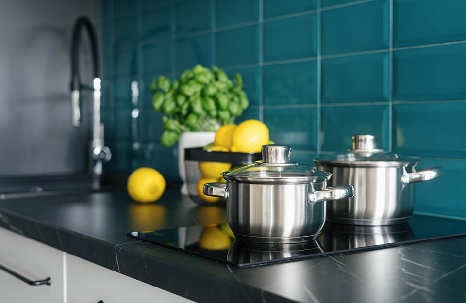 Primo piano sui fornelli di una cucina con top in marmo scuro, piastrelle color ottanio e mobili bianchi, con limoni e basilico accanto al lavabo